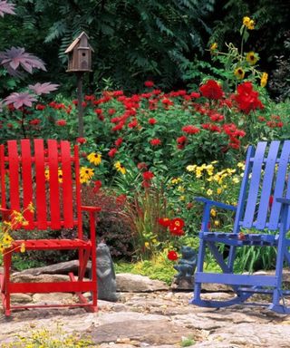 Two deck chairs - one red and one blue - on a stone patio area with red and yellow flower bushes behind them