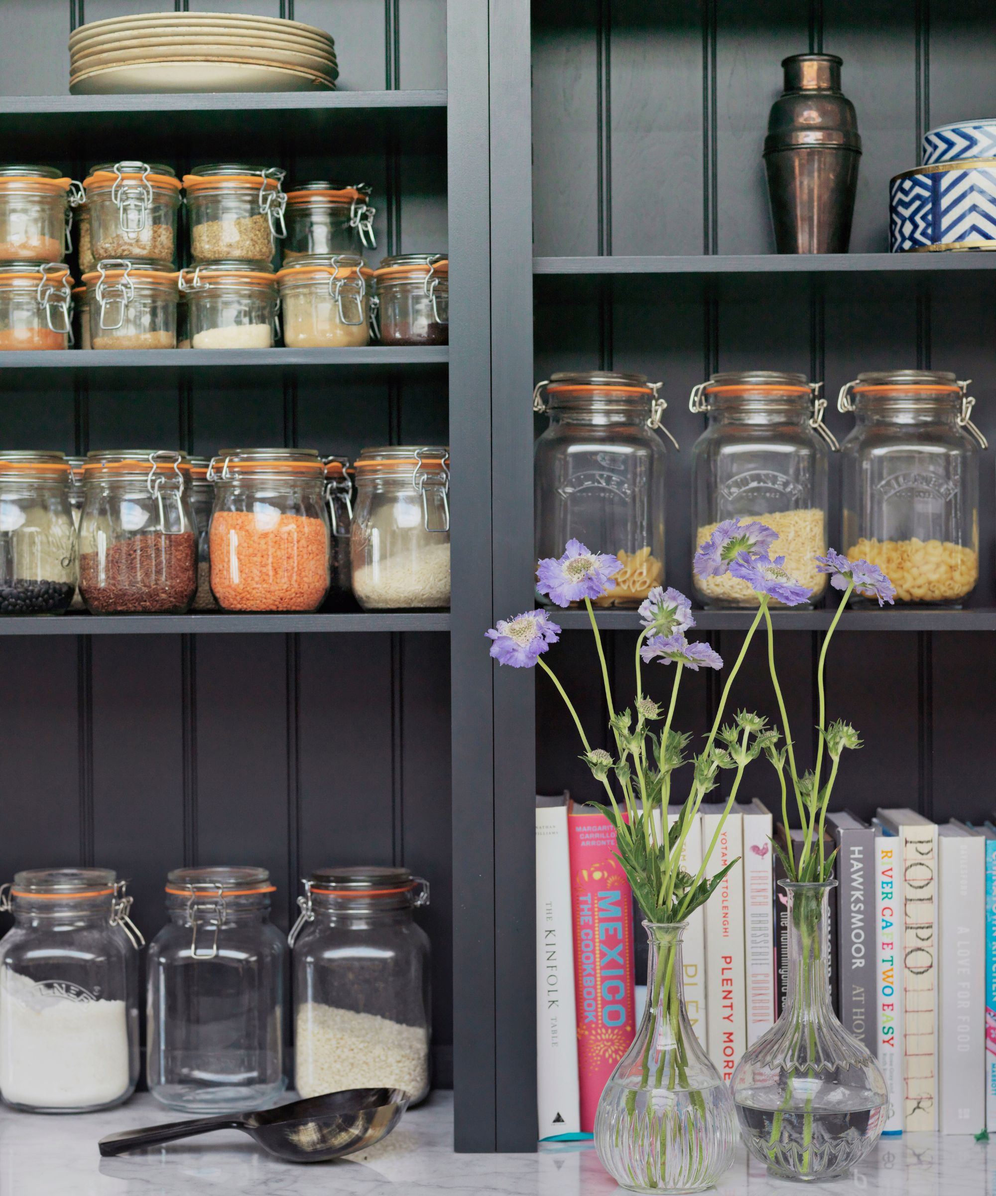 A pantry with black shelving, glass food storage jars, recipe books and two glass vases filled with purple flowers.
