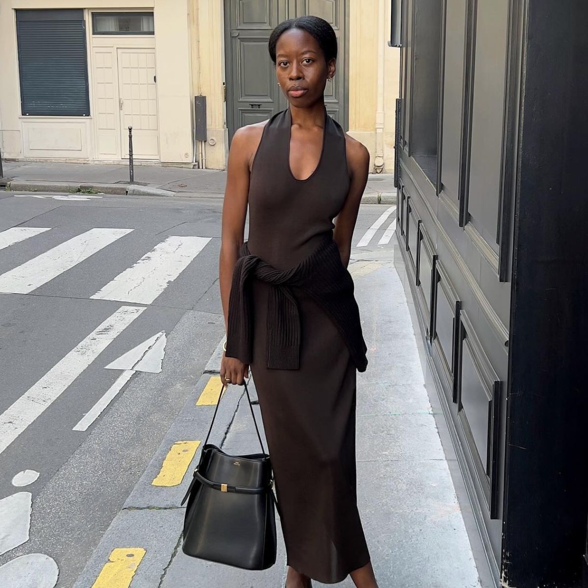 Woman wears brown halterneck dress, black jumper tied around the waist and carries black toteme bag