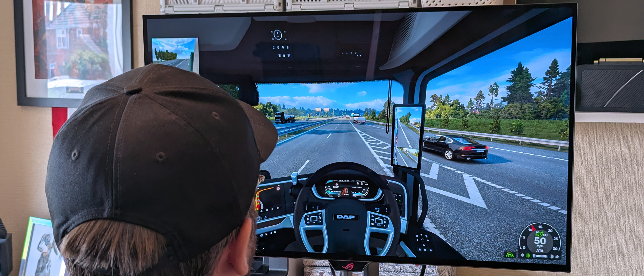 Windows Central's Ben Wilson wearing a black cap sits at a desk playing Euro Truck Simulator 2, with the ASUS ROG Strix OLED XG32UQWMS monitor showing a DAF truck interior.