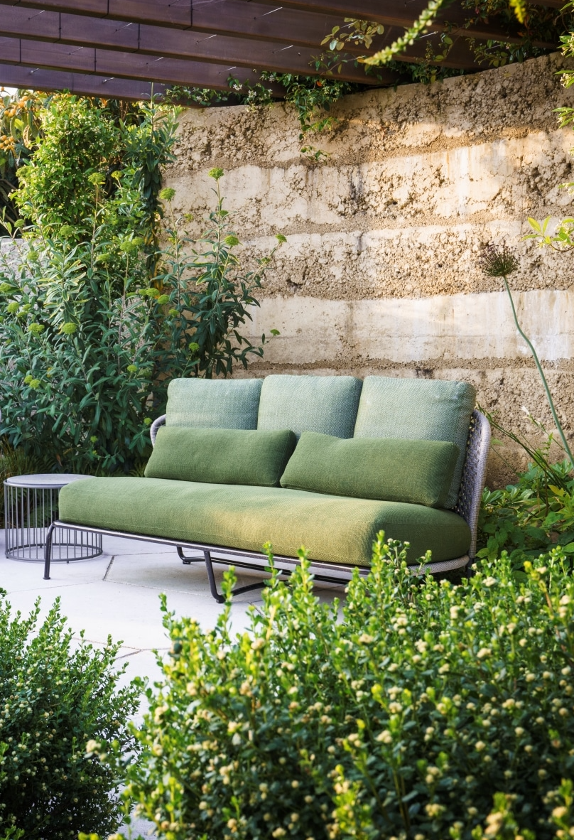 Green outdoor sofa with two-tone cushions and woven seat against a modern-rustic stone wall