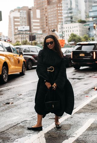 One of the best designer belts is shown in a close street-style photo of a woman wearing a black headband, black sunglasses, a black plissé dress with a black Bottega Veneta belt, a black bag, and black tabi-toe flats