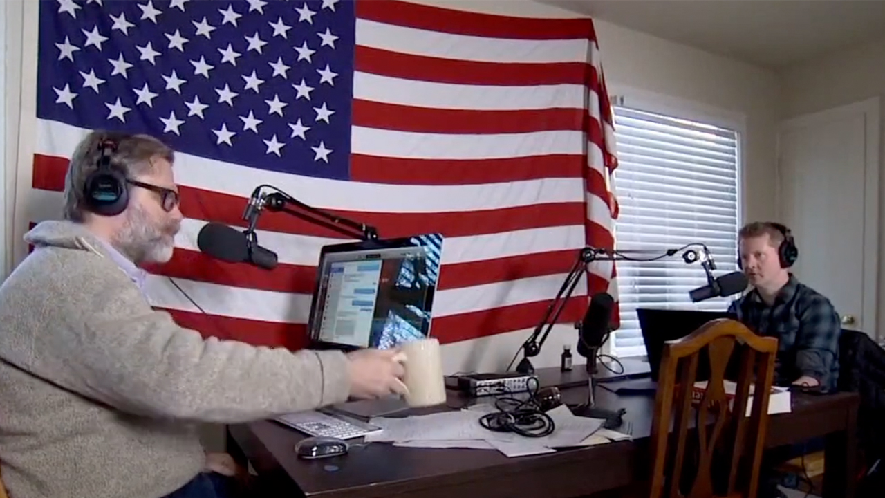John Roderick in front of an American flag on the left, sitting at a computer across from Ken Jennings while recording Omnibus