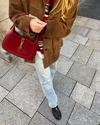 French style influencer Sabina Socol poses on a Paris sidewalk wearing a Reformation brown suede jacket, red striped sweater, light straight-leg jeans, black loafers, and a red Gucci Jackie bag.