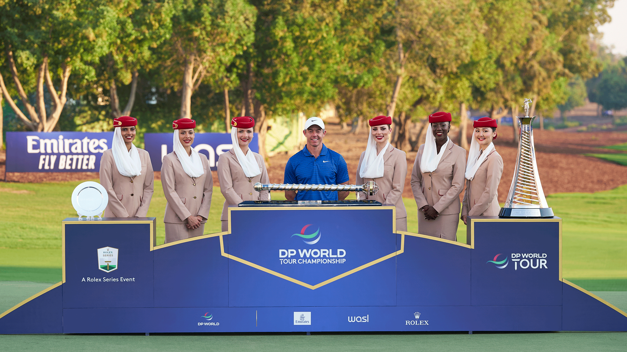 Rory McIlroy stands behind the DP World Tour Championship and the Race To Dubai trophies with six Fly Emirates flight attendants alongside him in 2024