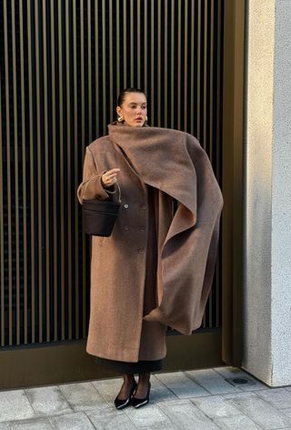 A woman wearing a long brown coat with an attached long scarf styled with gold earrings, a black bucket bag, a black skirt, black tights, and black kitten heel pumps.