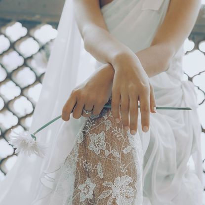 A bride in a delicate lacy wedding dress, holding a white flower and wearing a simple French manicure