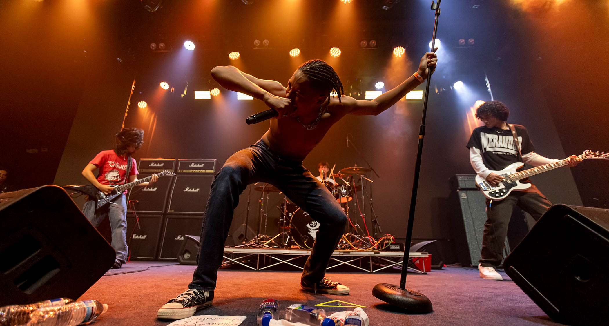 Chained Saint throw it down live with frontman Sean Sterling screaming into the mic, guitarist Ethan Kahn wearing a red T-shirt and playing a black Jackson Rhoads, and bassist Sebastian De Avila playing a Jackson Concert bass guitar.