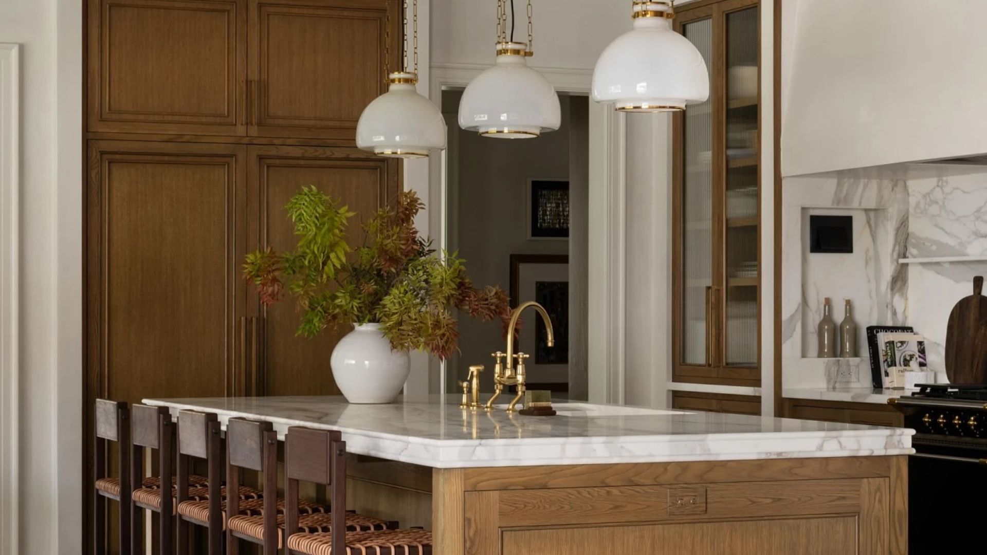 A kitchen with wooden cabinets, a wood island with a stone top, a row of island stools, a vase of leafy stems, gold hardware, and three overhead pendant lamps