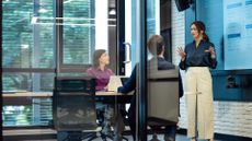 Female business leader standing in a meeting room discussing company strategy with male and female colleague, with statistics on screen behind her. 