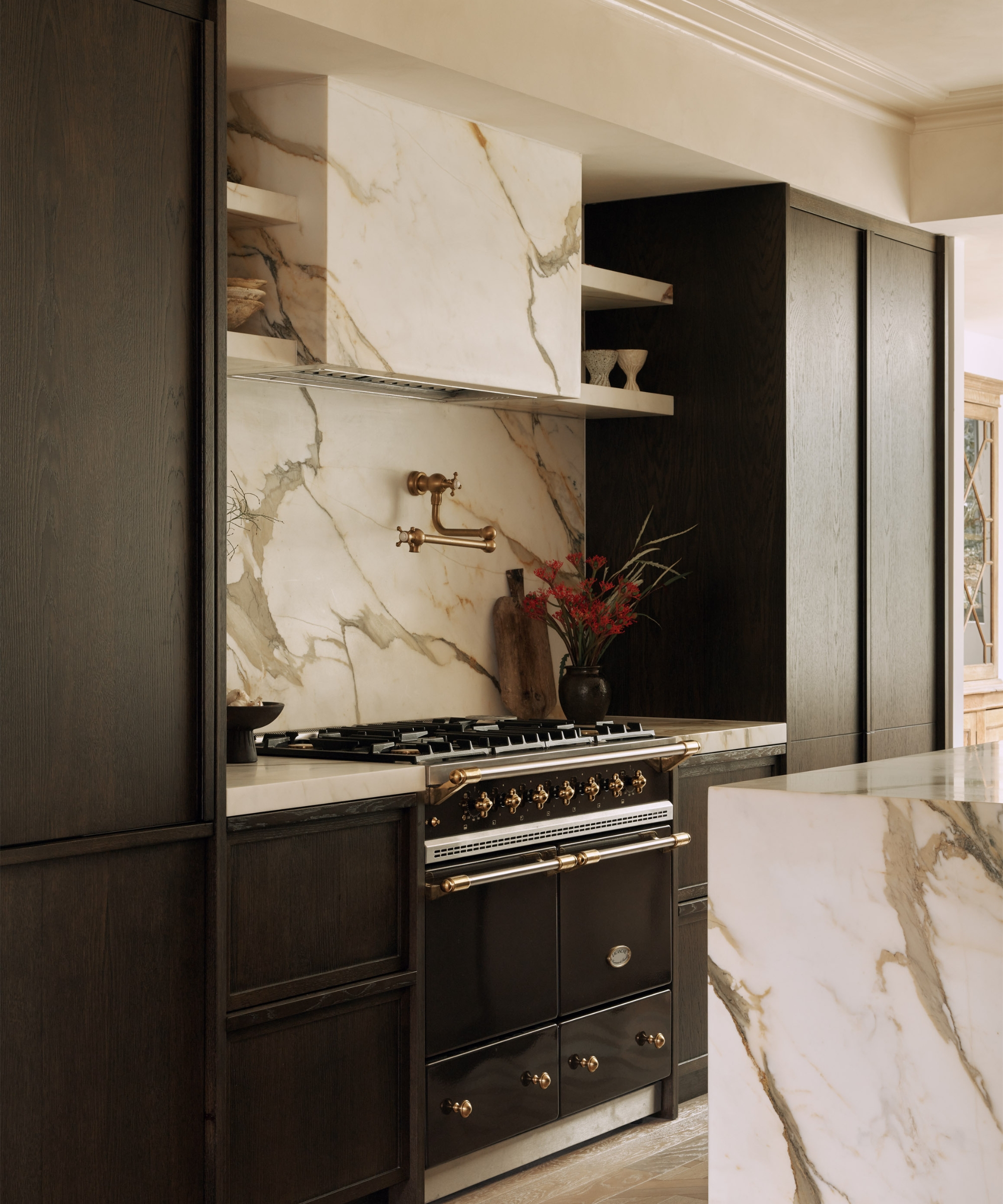 A moody kitchen with blackened wood cabinets, beige marble for the counters and backsplash, and a black range with brass accents