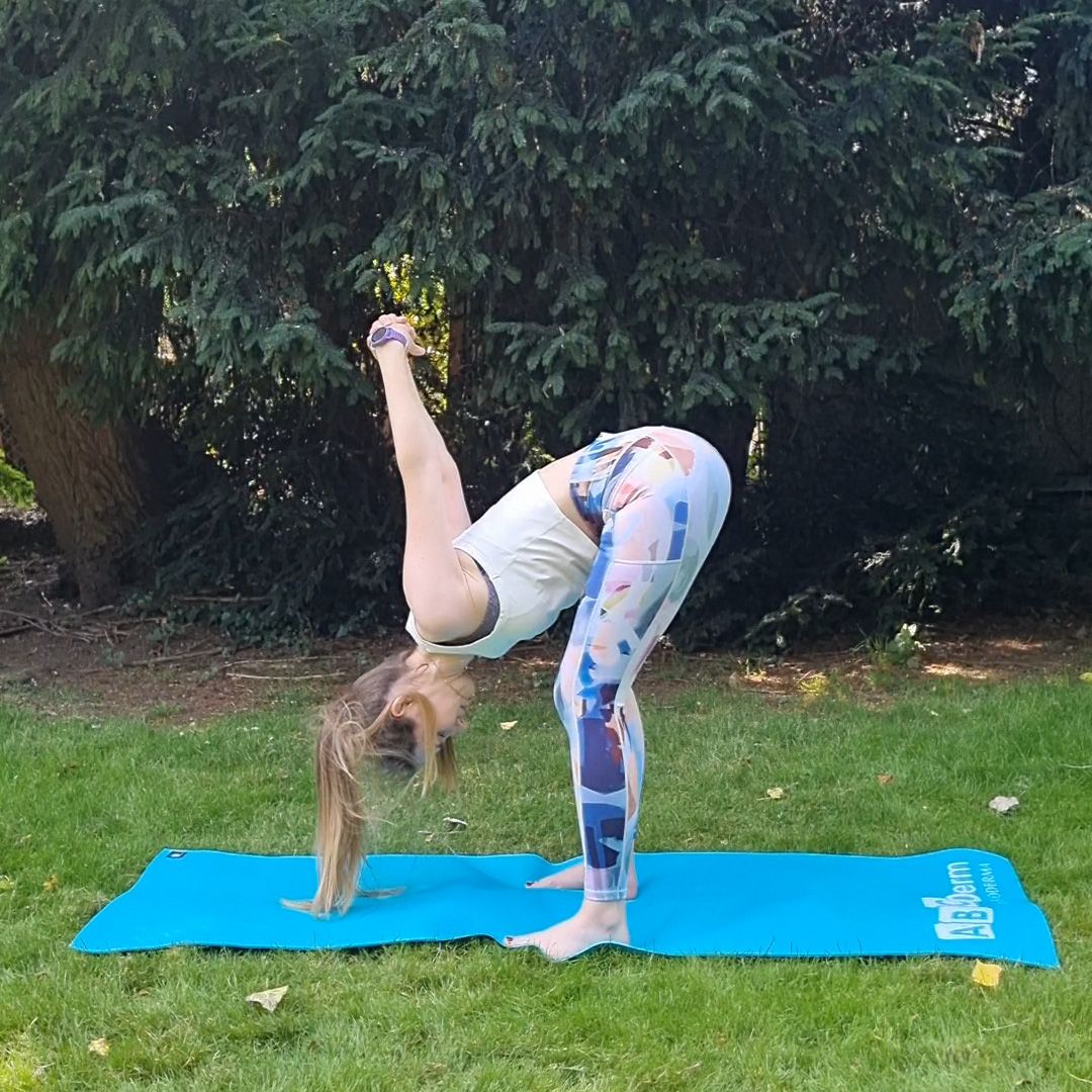 Fitness editor Ruth Gaukrodger performs a shoulder and chest stretch to forward fold on a yoga mat in a green outdoor setting. She is standing but hinging at the hips to perform a forward fold, with her hands clasped behind her and pointed to the sky. Her head is close to her calves.