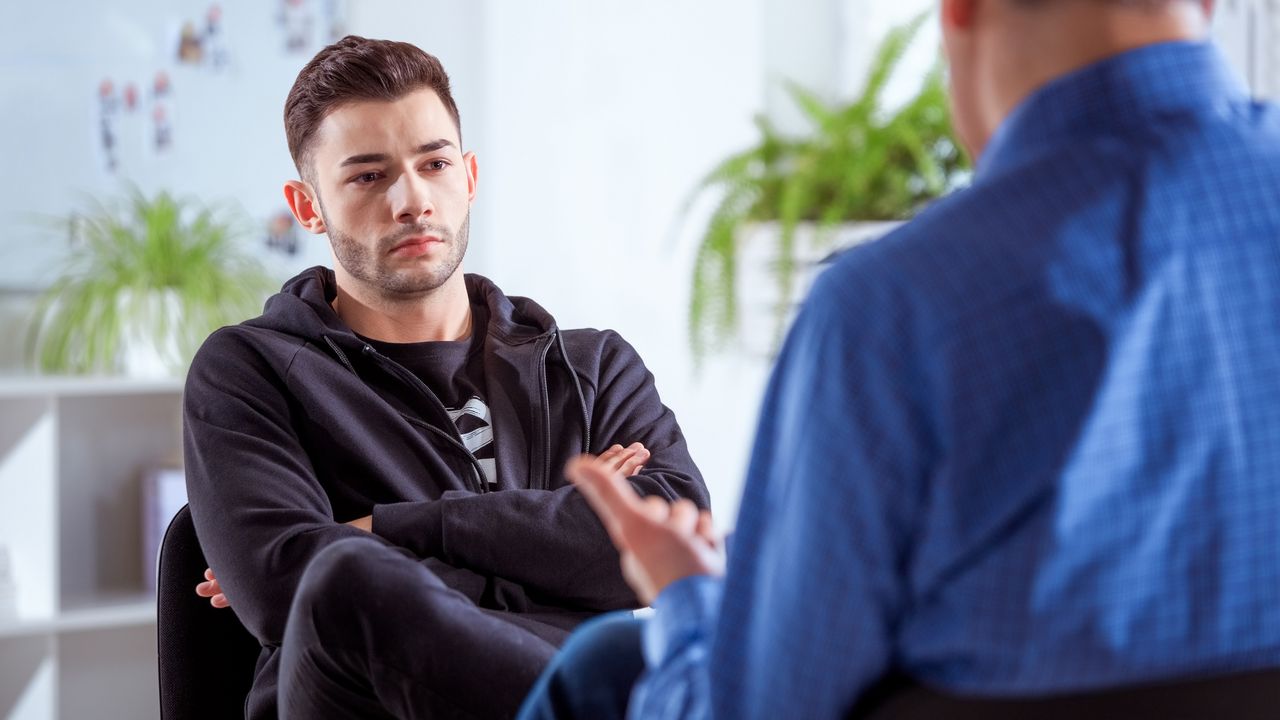 A young man sits with his arms crossed while an older man talks to him in an office setting.