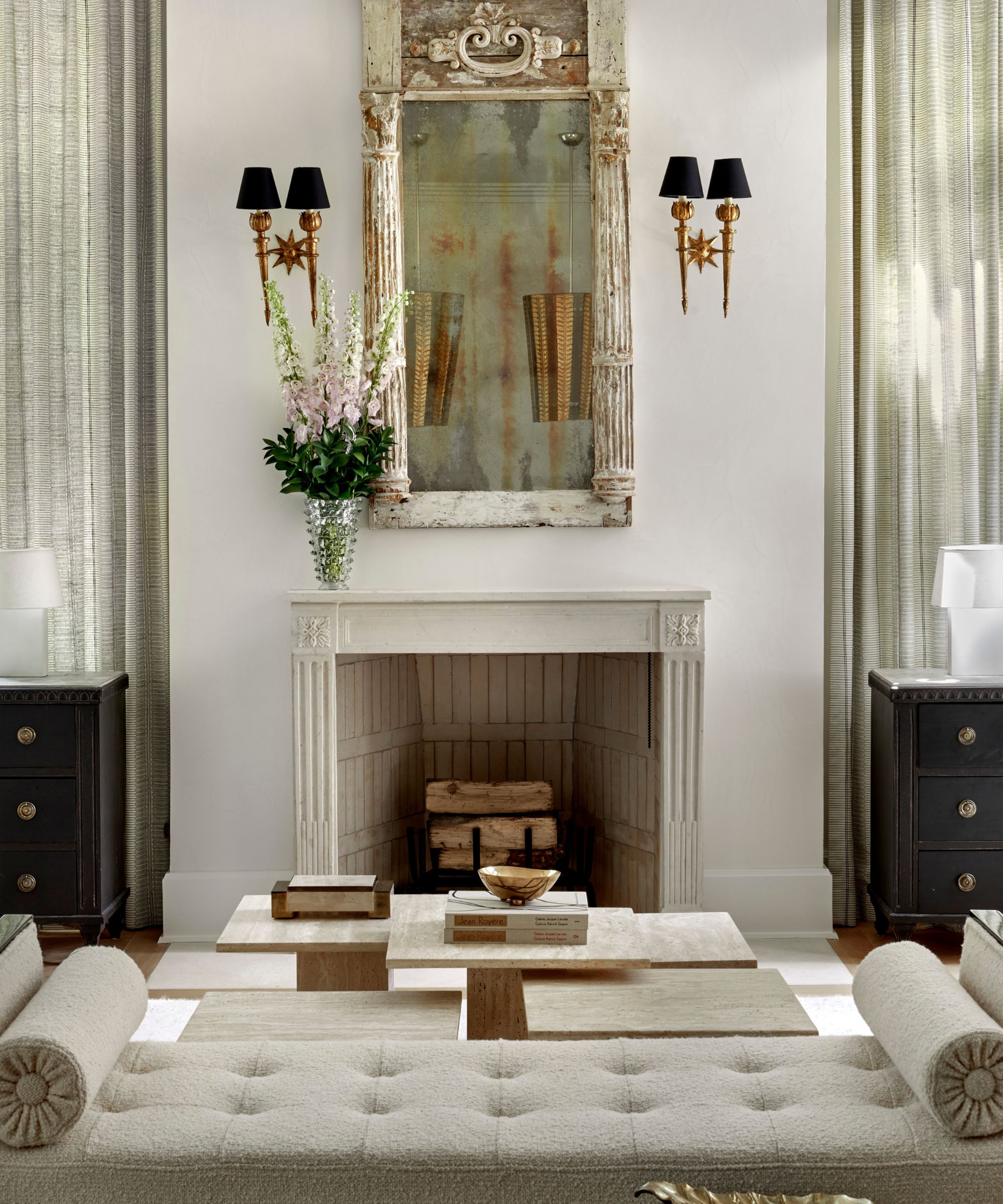 A formal living room featuring a white carved stone fireplace, a tall weathered antique mirror, and symmetrical gold wall sconces with black shades.