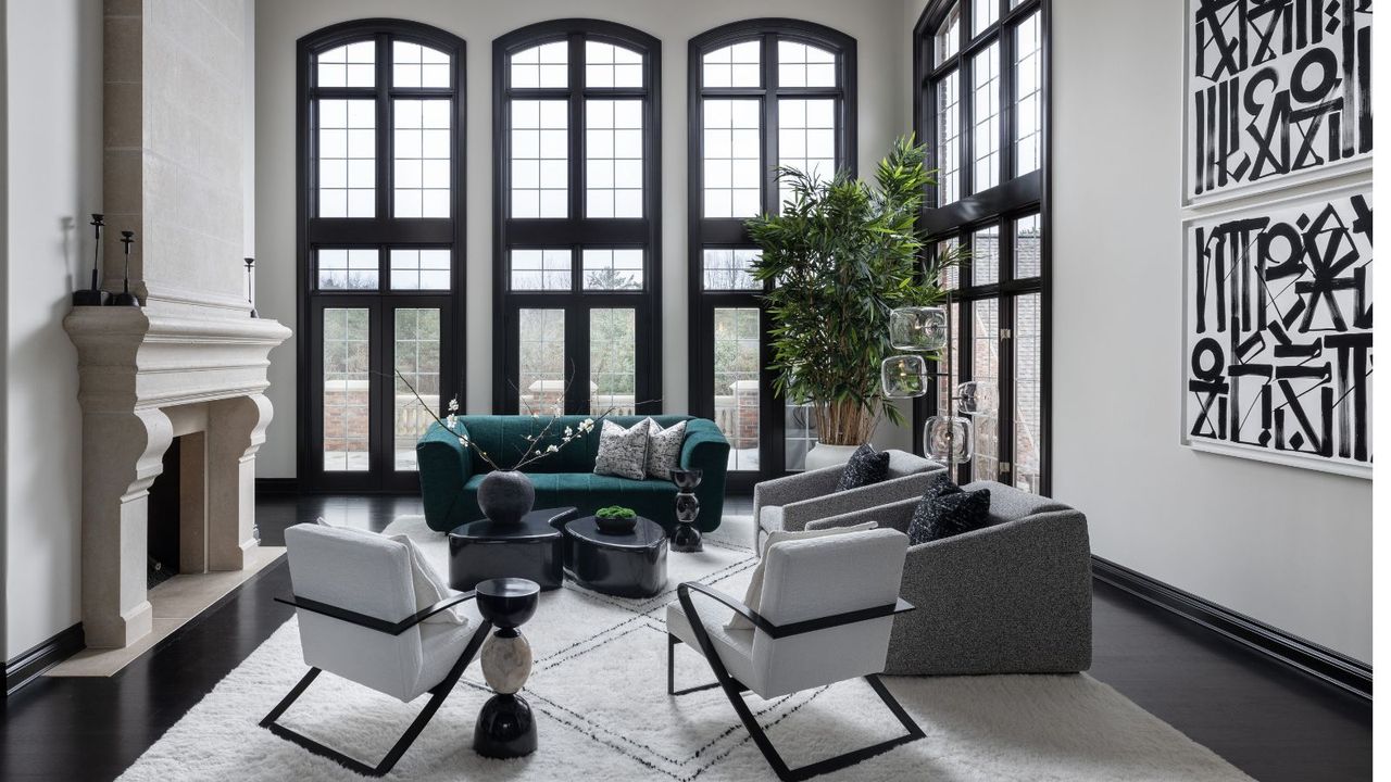 Large high ceiling living room in black and white with black framed floor to ceiling windows and featuring an accent sofa in dark green. There is a large cream stone fireplace on left and black and white abstract artwork on right wall