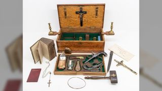 This is an image of a vampire slaying kit. It contains a lot of items, such as a bible, rosary beads, several flasks of holy water, a pair of pistols, a crucifix, a plain communion book, a wooden hammer, and a necklace of sorts.