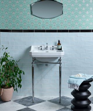 A bathroom with seaform green wallpaper with a gray mirror on it, light blue tiles with a white standing sink in front of it, and a green plant, black vase with a towel on and diamond tiles on the floor