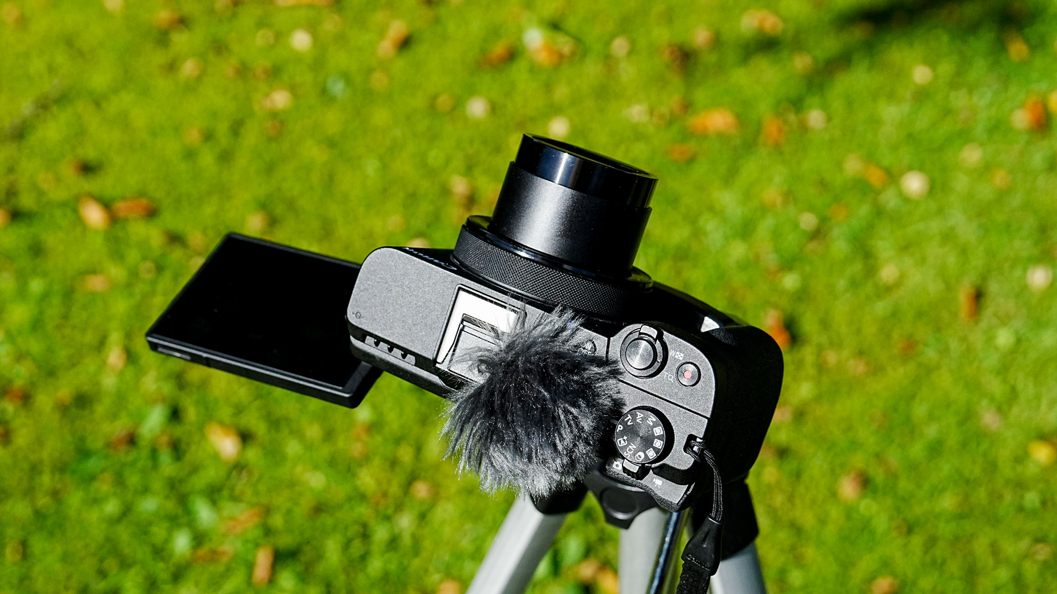 A Canon Powershot V1 with shutter open on a tripod with grass in the background.