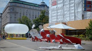A wide-angle shot of a public plaza in Vienna, Austria, showcasing a street art installation on a sunny day. In the foreground, a large abstract sculpture made of a twisted metal frame and various reflective geometric shapes, including pyramids, sits on a circular concrete base. 