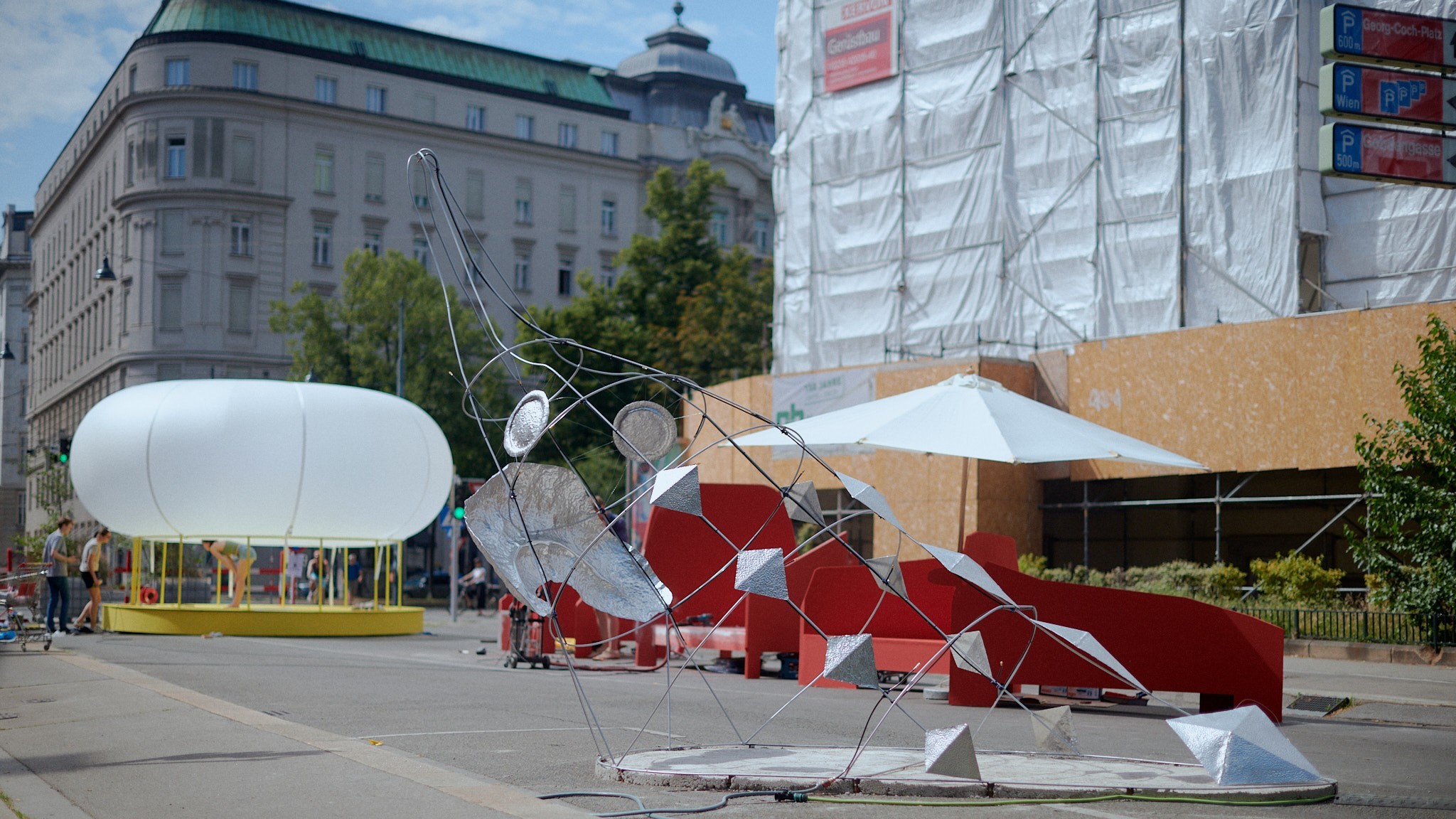 A wide-angle shot of a public plaza in Vienna, Austria, showcasing a street art installation on a sunny day. In the foreground, a large abstract sculpture made of a twisted metal frame and various reflective geometric shapes, including pyramids, sits on a circular concrete base.