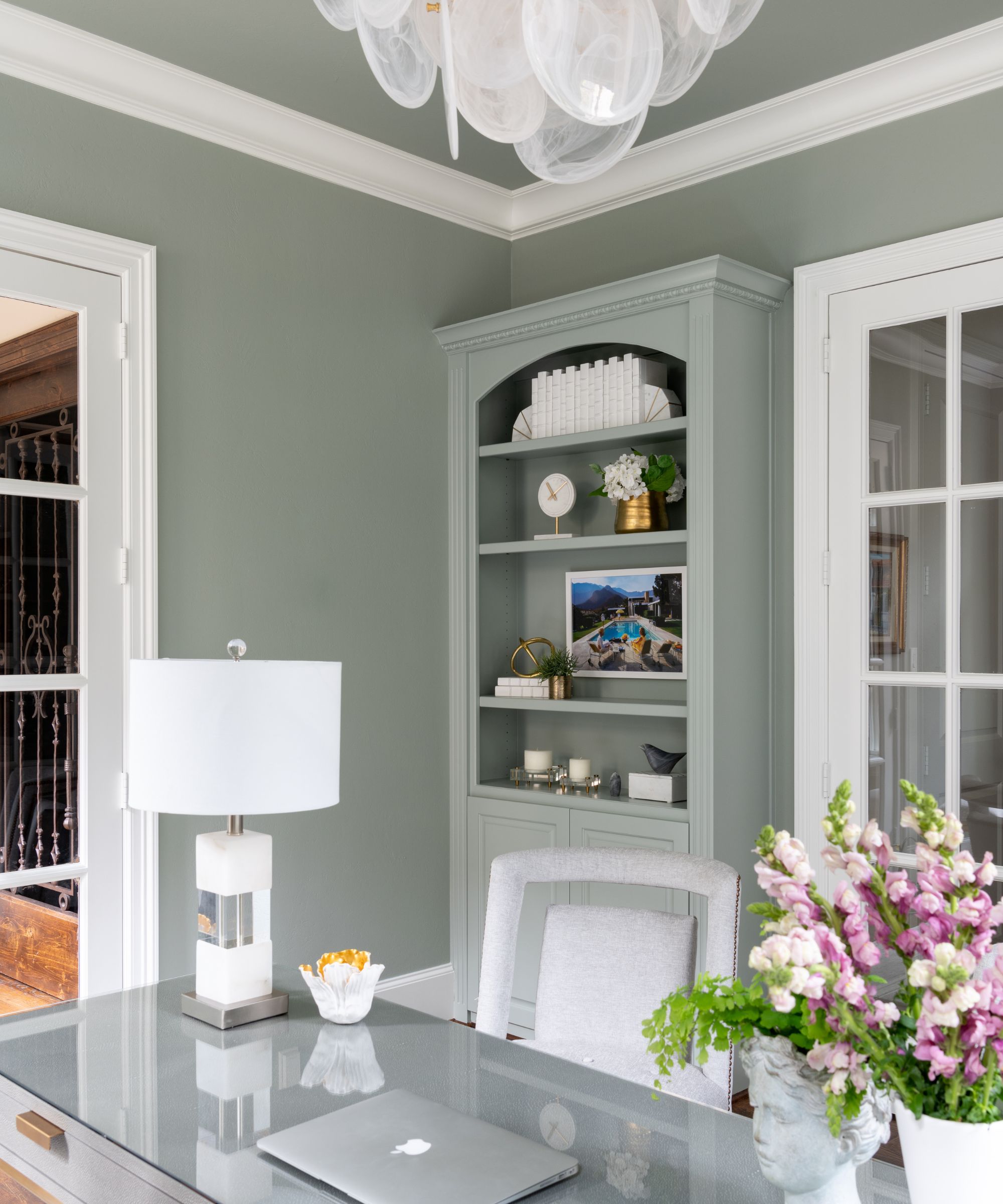 Home office painted in Sherwin-Williams Oyster Bay, a desk with a velvet desk chair and white lamp, a built-in bookcase with pictures and decor, and a vase of foxgloves.