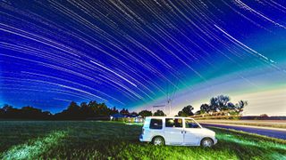 a white van is parked in a grassy field next to a road, lined with the streaking lights of a passing car. the sky is filled with the trails of stars.