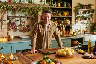 Jamie's Easy Christmas: Jamie Oliver stands in his kitchen, resting his hands on the countertop in front of him, smiling at the camera. There is a chopping board with a knife next to it on the counter in front of him, and a dish full of peeled potatoes next to it. The kitchen is festooned with burning candles.