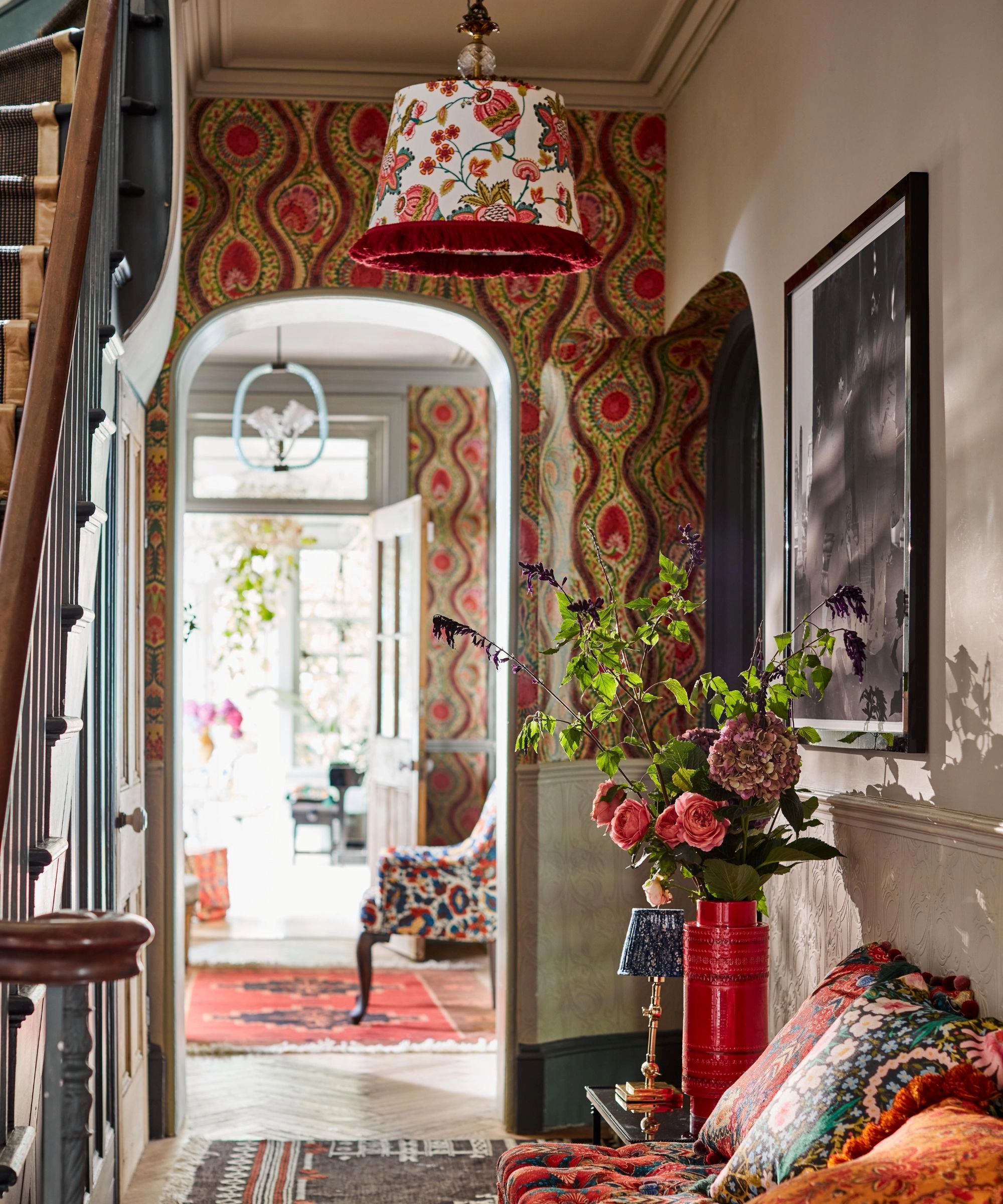 maximalist entryway with floral wallpaper, floral ceiling light, colorful rugs and colorful staircase with a patterned bench seat and patterned pillows