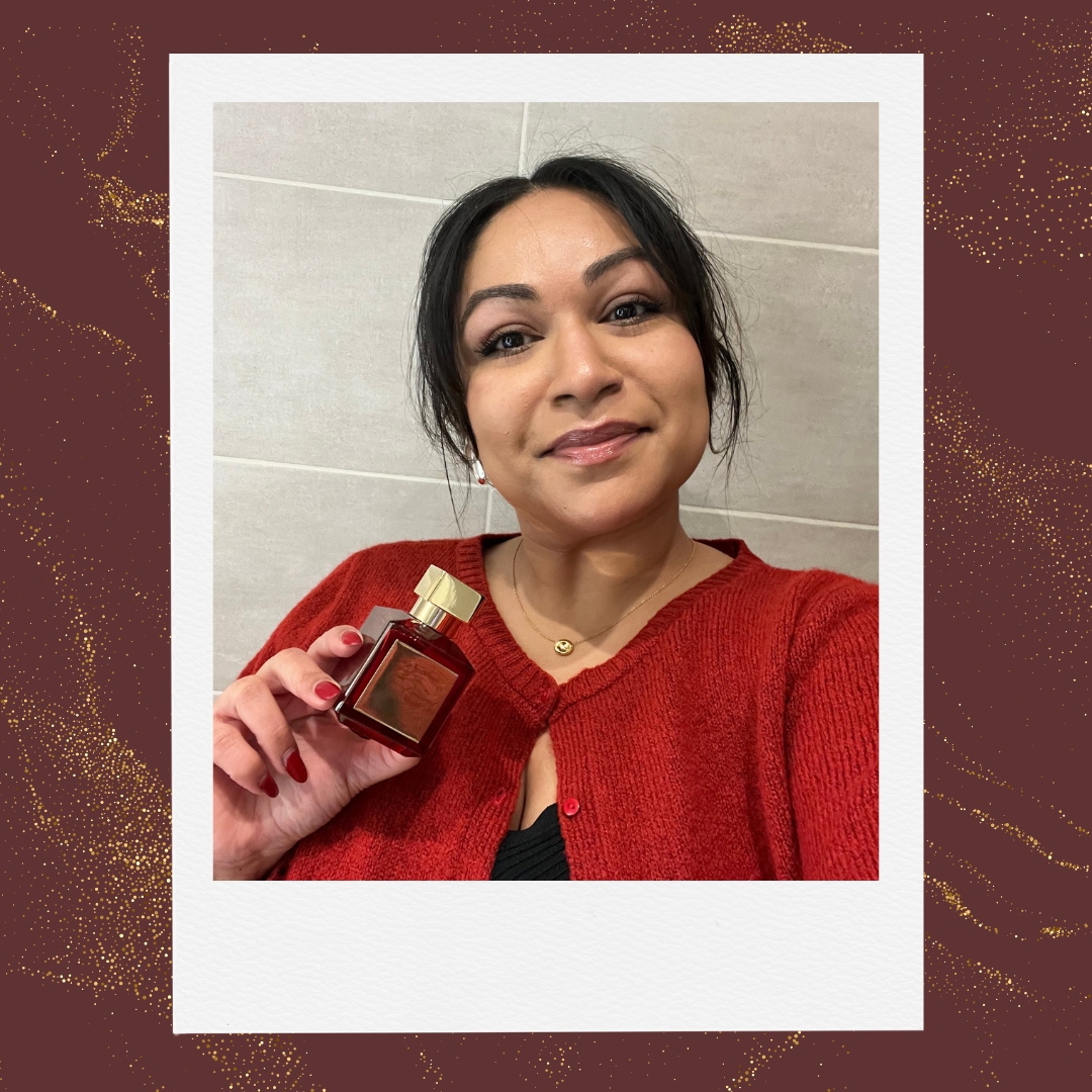 A picture of Digital Beauty eCommerce Editor, Aleesha Badkar, seen wearing a red cardigan and holding a bottle of the Maison Francis Kurkdjian Baccarat Rouge 540 Extrait de Parfum, which is red and gold/ in a burgundy template with a gold glitter overlay