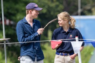 Lady Louise Windsor and Felix da Silva-Clamp wearing blue shirts and smiling outdoors in front of a rope fence with red and white bunting as Felix taps Louise with a feather