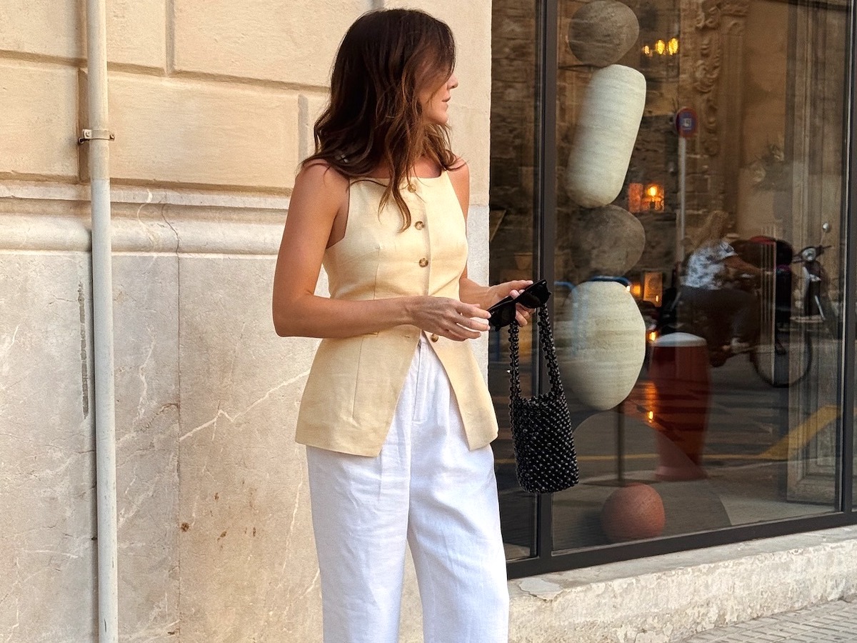 British influencer Kim Turkington poses on a sidewalk in Palma De Mallorca, Spain wearing a butter-yellow button-front vest, a black beaded mini bag, and white linen pants