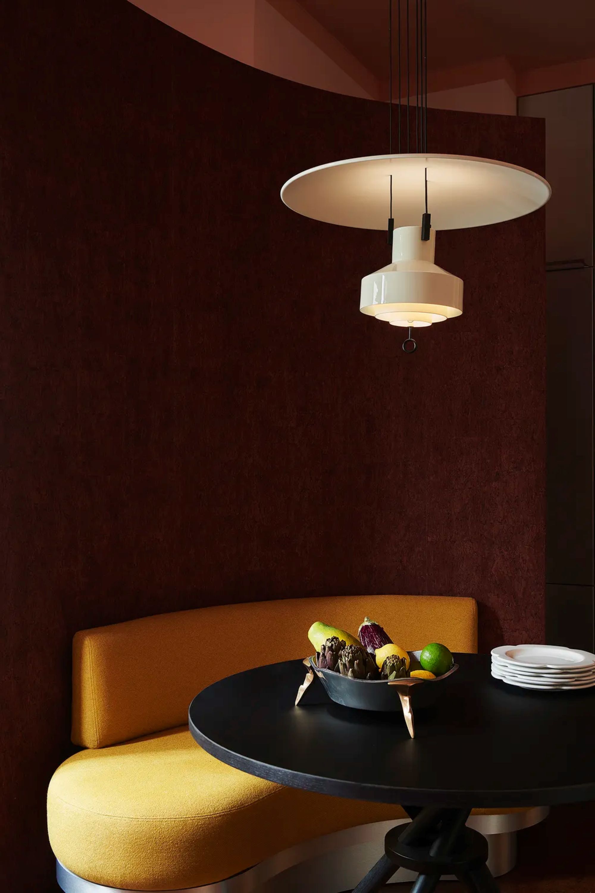 A small dining space with a curved, yellow sofa, a black, round table, a bowl of fruit, a stack of white plates, and a designer pendant light