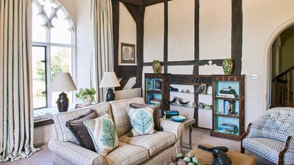 Cream Tudor living room with high ceilings, dark wooden beams on the far wall and tall arched windows with ornate metal work. The beamed wall has built in alcove shelving, and the sofas are cream and mismatched but cozy