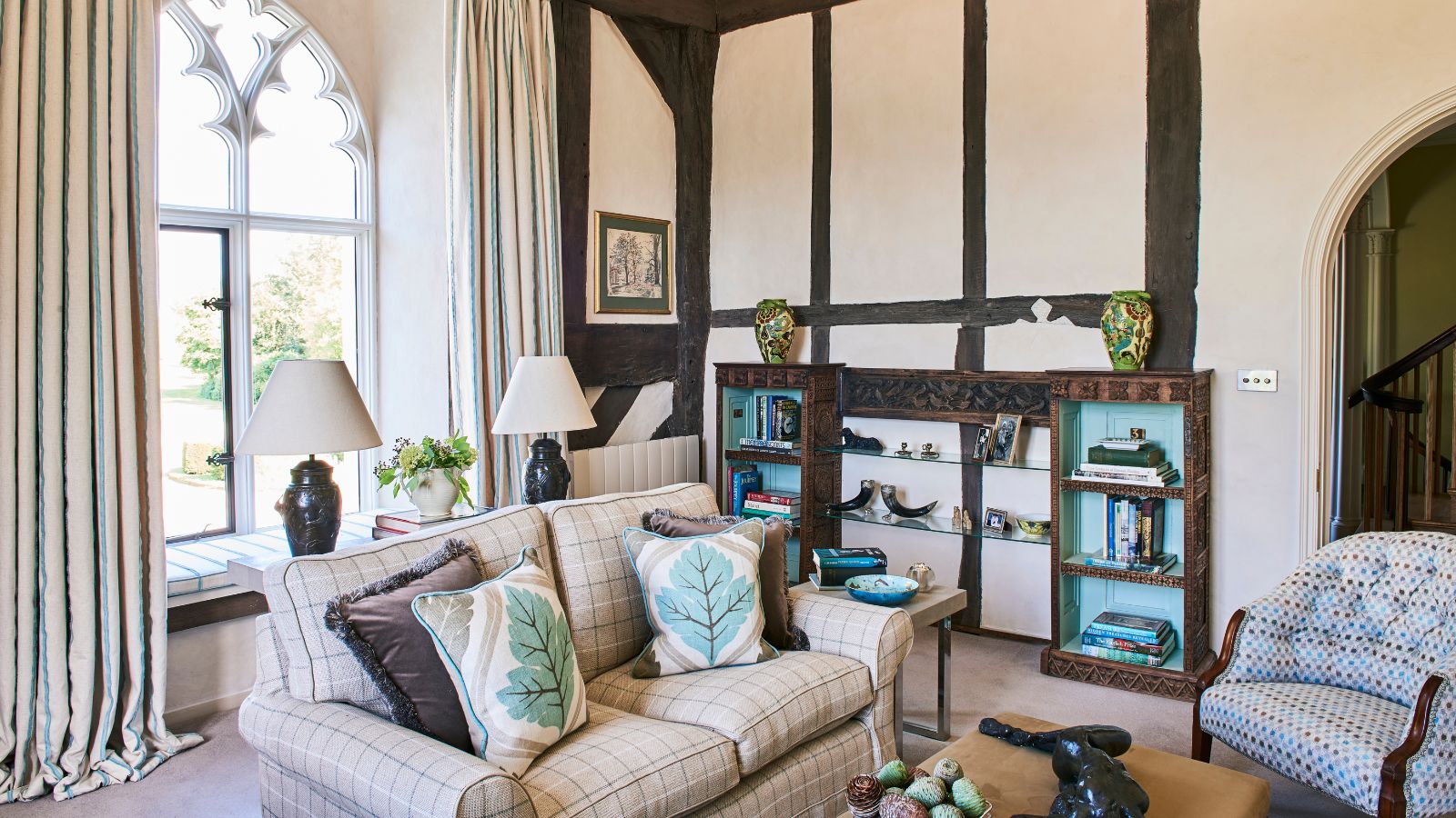 Cream Tudor living room with high ceilings, dark wooden beams on the far wall and tall arched windows with ornate metal work. The beamed wall has built in alcove shelving, and the sofas are cream and mismatched but cozy