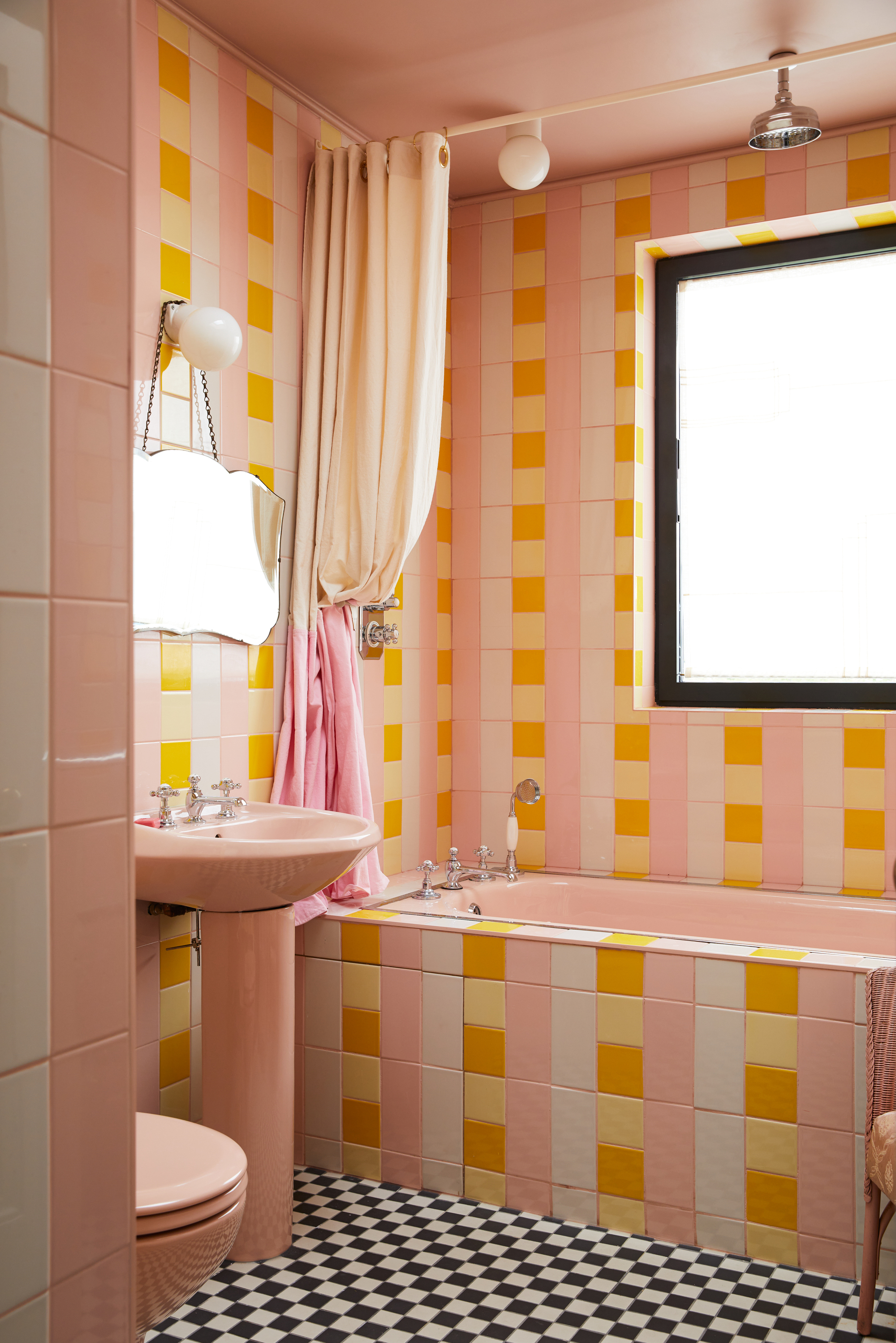 a colorful bathroom with yellow, pink, and white tiles laid in a plaid pattern, with black and white checkered tiles on the floor, a pink pedestal toilet and sink with a shower curtain and antique mirror