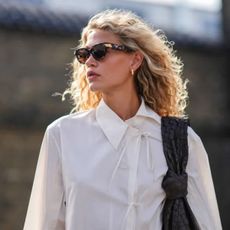 a woman with beautiful blonde wavy hair walks down the street wearing a white shirt and sunglasses
