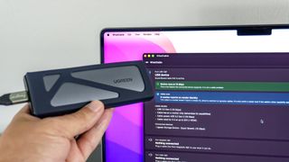 A person holding up a portable SSD in front of a MacBook Pro running the WhatCable app to test the speed of the cable connecting the two