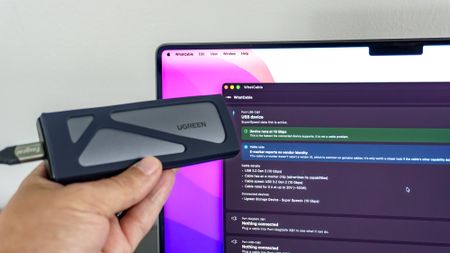 A person holding up a portable SSD in front of a MacBook Pro running the WhatCable app to test the speed of the cable connecting the two