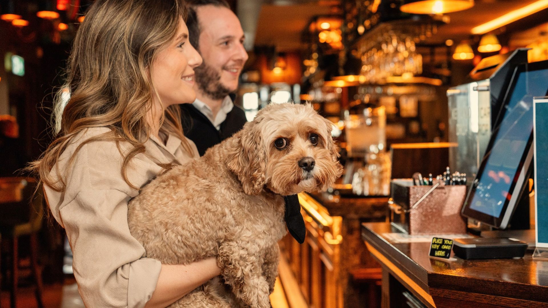 A couple enjoy drinks at the cosy bar of a dog-friendly inn, cuddling their dog as they order.