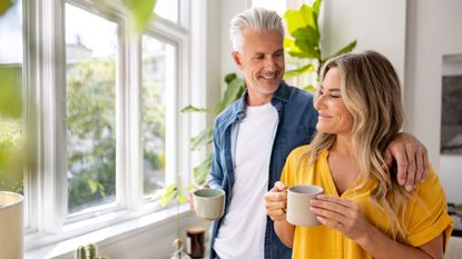 A Gen X couple smile as they look out the window of their kitchen while holding coffee cups.