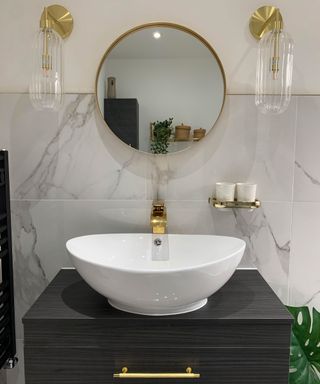 Round mirror with gilded frame hung over white basin in gray marble effect bathroom