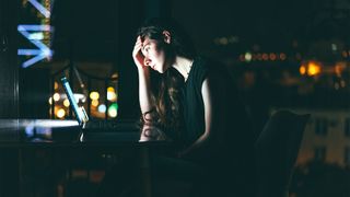 Female IT worker dealing with tech overload while working in a dark room with face illuminated by computer screen.