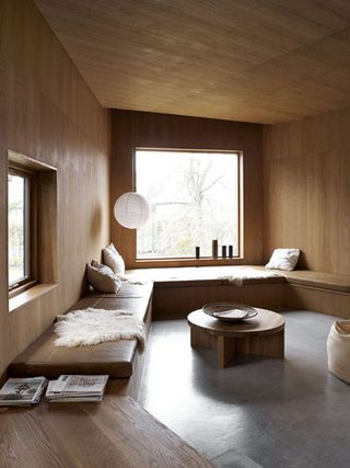 The room has concrete floors, wood-clad walls, and a sitting area that goes all around the room and is covered with brown leather furnishings. Large windows cover the walls, one on each side.