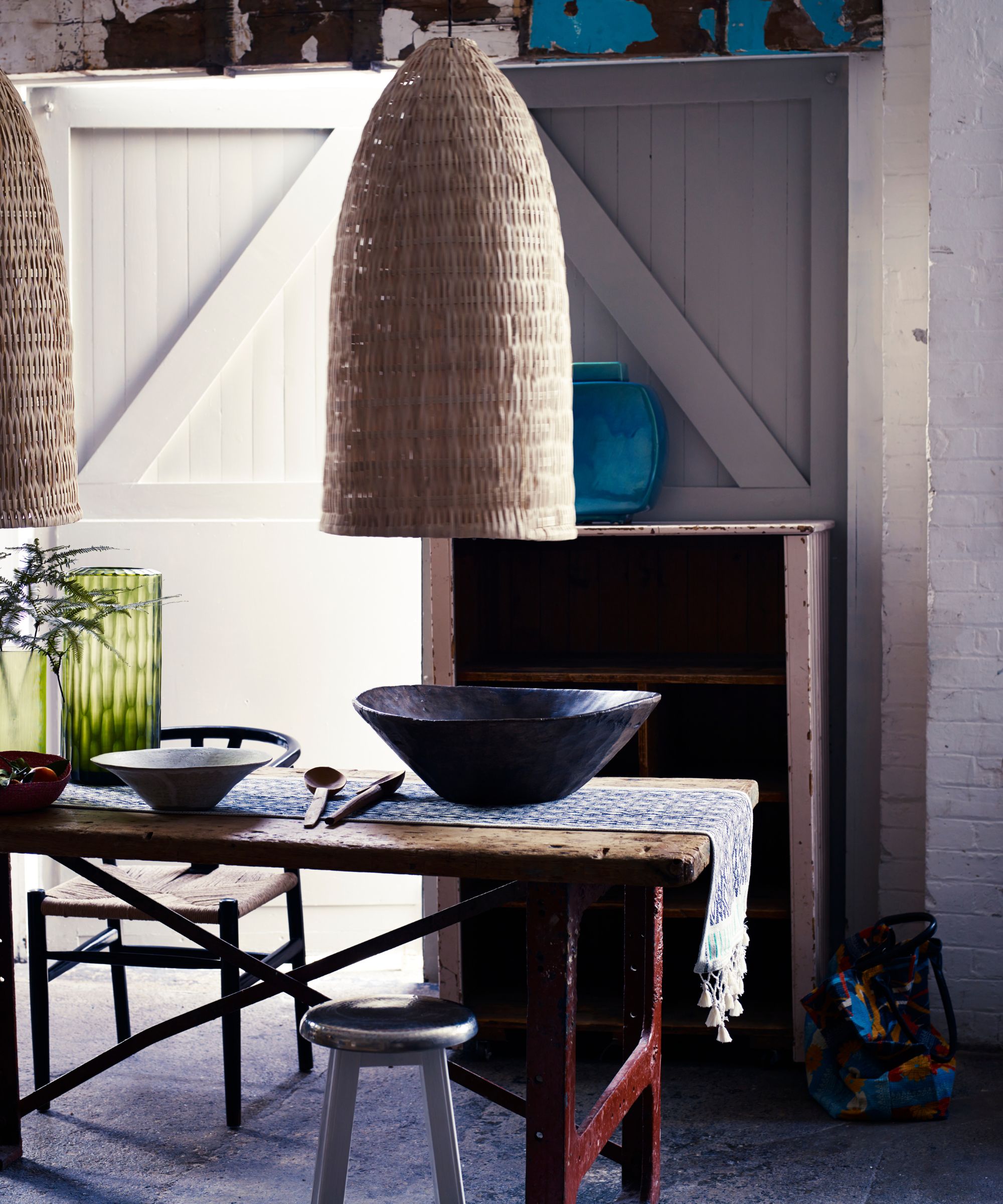 A dining room with long wooden table and stools, decorative bowls and a patterned table runner, and two large rattan lightshades hanging above. Behind the table is an empty pink shabby chic style bookshelf with a blue vase on top, and behind are two panelled doors. The left door is open, and letting light into the room.