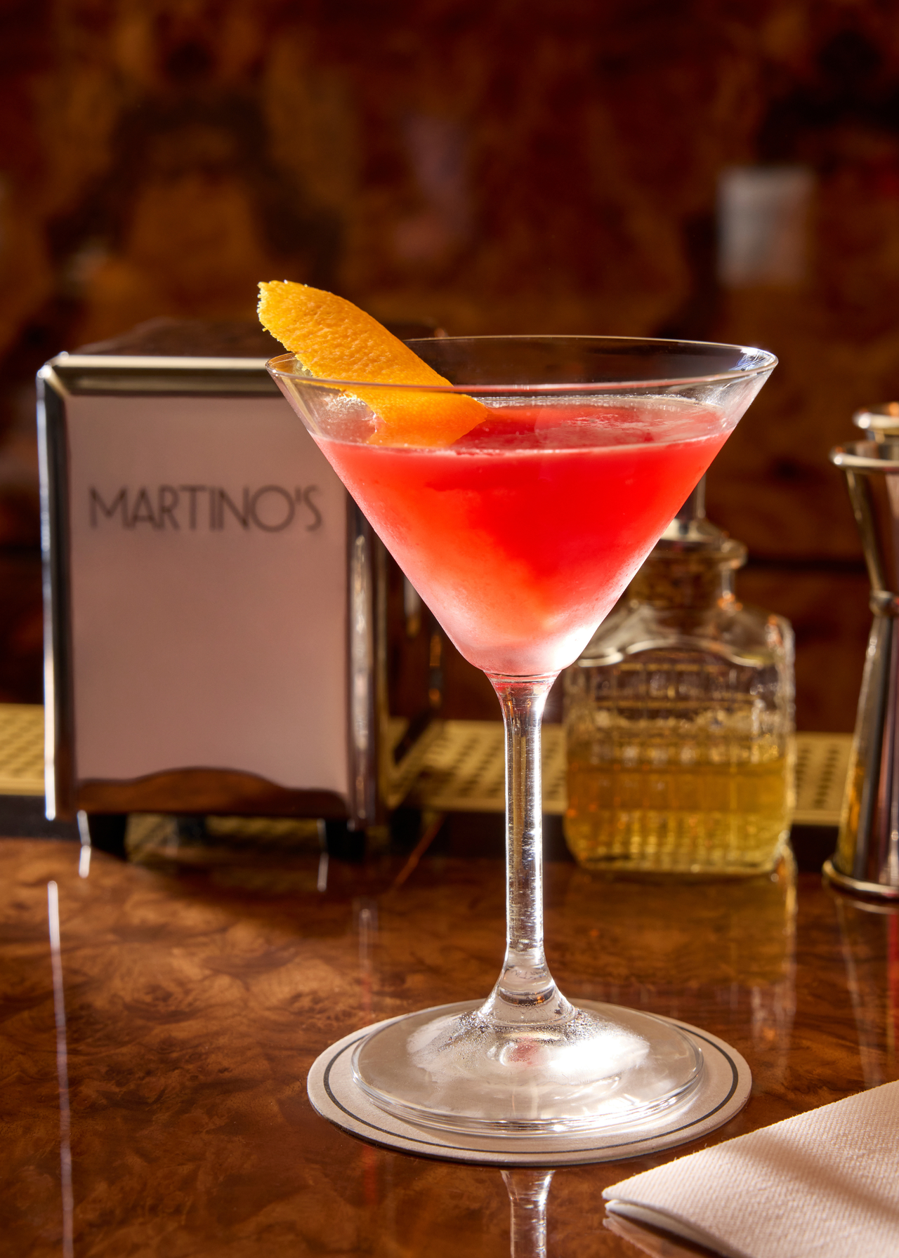 a red cocktail served in a martini glass, garnished with orange peel