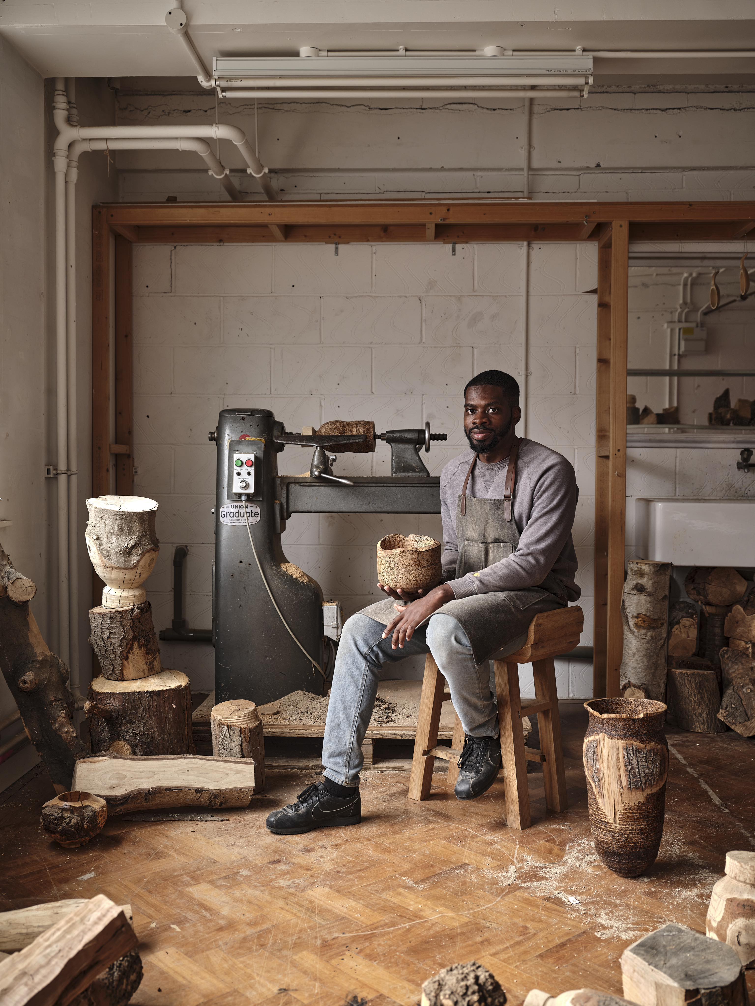 Artist, designer and craftsman Darren Appiagyei, sits on a stool in workshop next to lathe surrounded by wooden sculptures