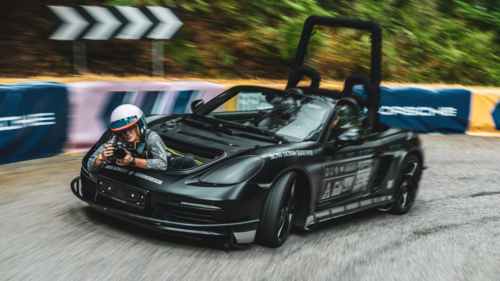 A modified black Porsche convertible, featuring an exposed roll cage and a photographer leaning out of the hood compartment, is photographed driving quickly around a curve with event banners visible.