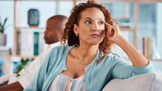 Mature woman on sofa thinking seriously, with man blurred in background