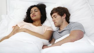 A woman with curly black hair sleeps next to a man with light brown hair, both covered by a white comforter