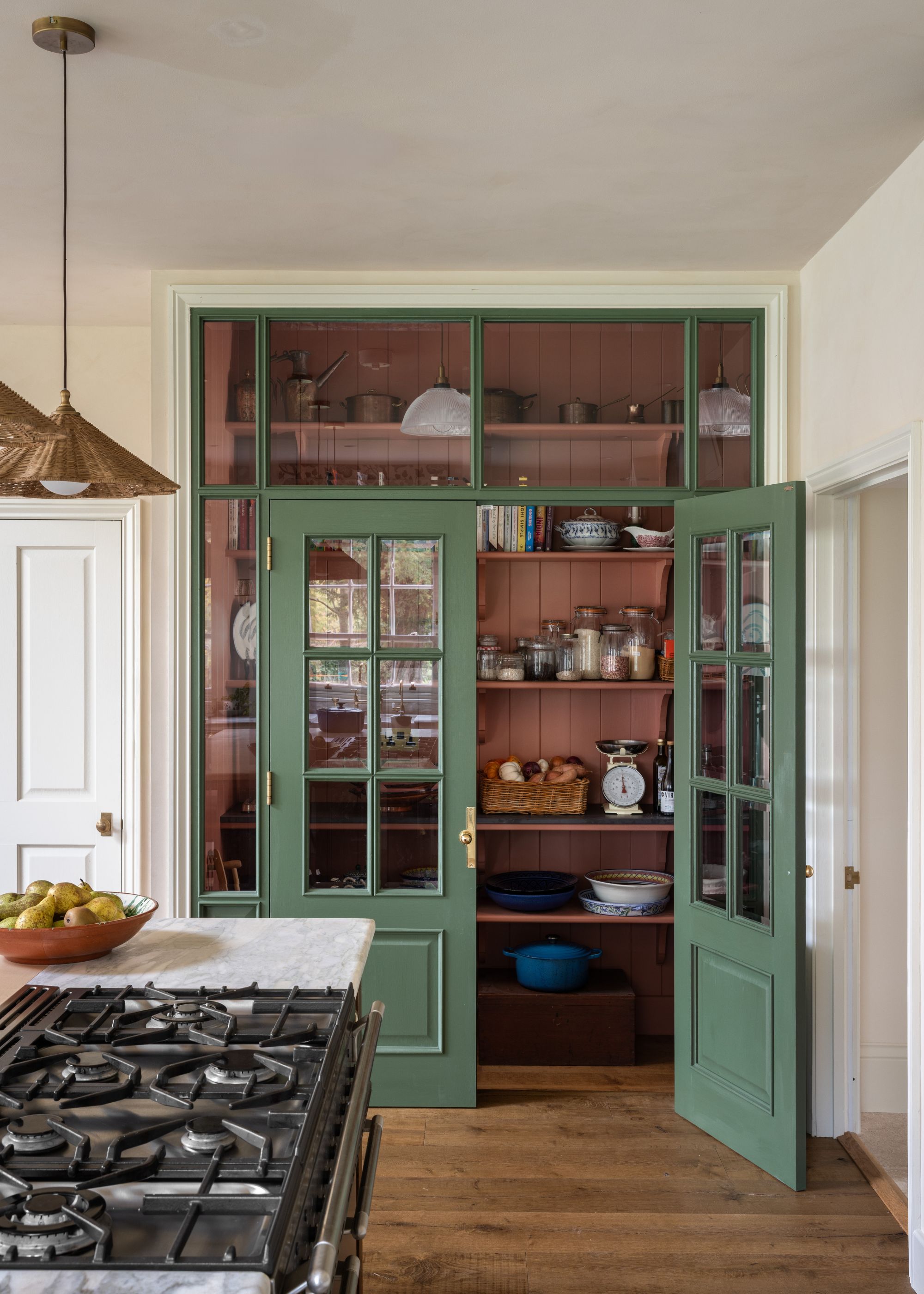 A large kitchen with off-white walls, wooden flooring, and a pantry with a dark, muted pink interior and dark green doors with glass panels.